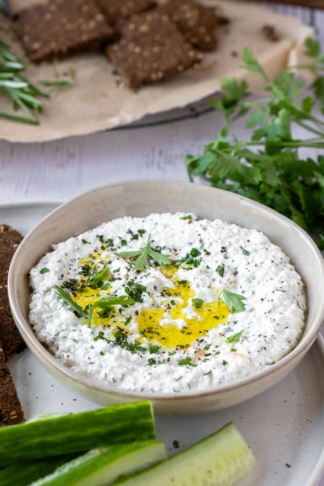 Romige cottage cheese dip met olijfolie, citroen en verse kruiden in een schaaltje, geserveerd met rauwkost en crackers — eiwitrijke, gezonde dip. Whipped cottage cheese.