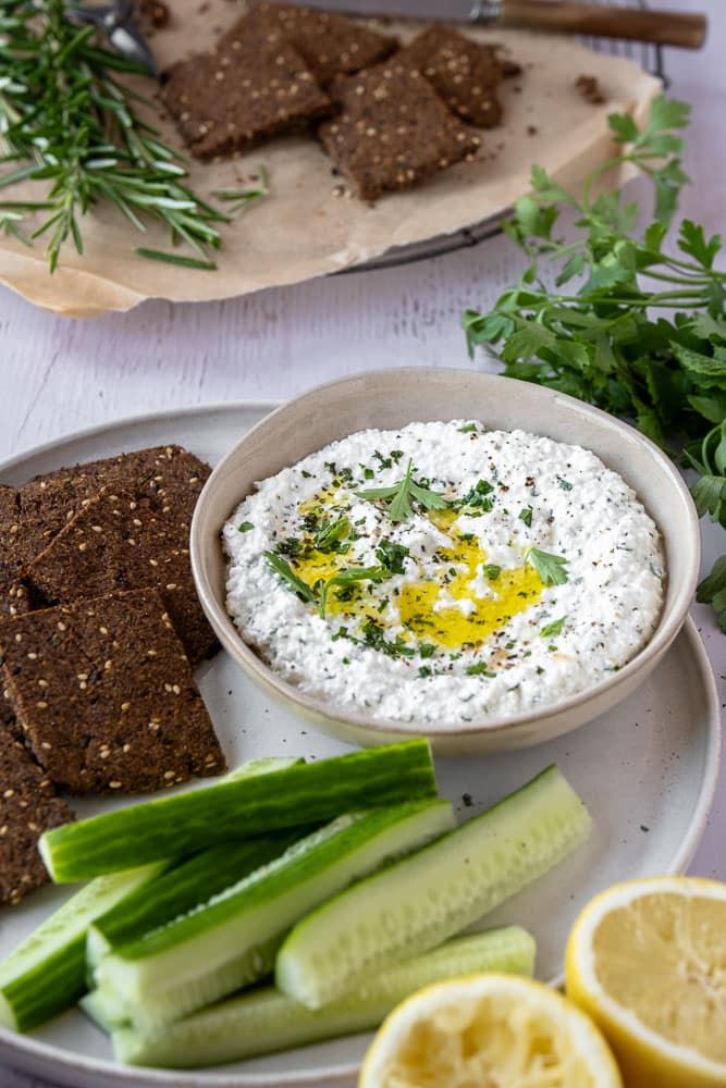 Romige cottage cheese dip met olijfolie, citroen en verse kruiden in een schaaltje, geserveerd met rauwkost en crackers — eiwitrijke, gezonde dip. Whipped cottage cheese.