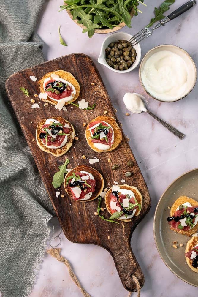 Blini’s met carpaccio, cottage cheese en rucola op kleine pannenkoekjes, geserveerd als feestelijk hapje.