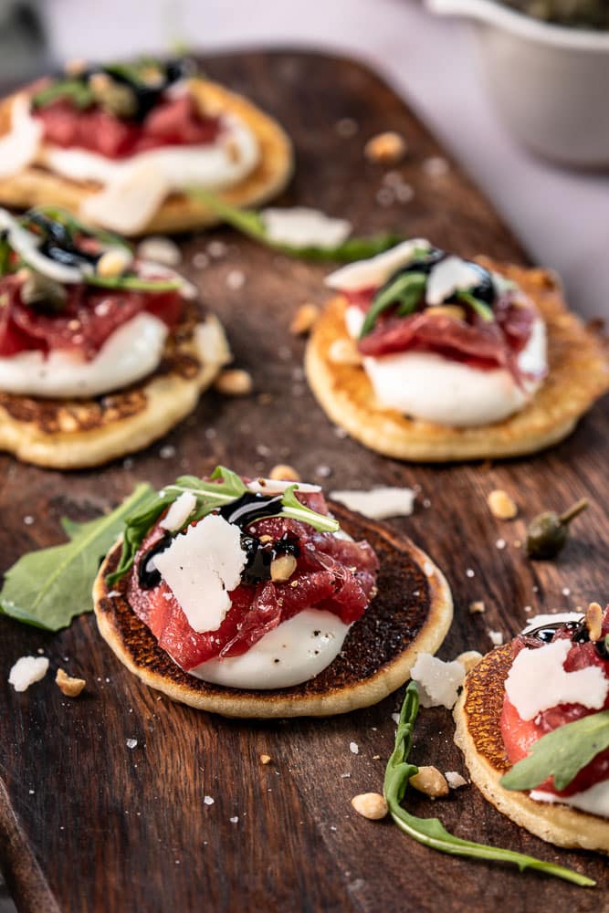 Blini’s met carpaccio, cottage cheese en rucola op kleine pannenkoekjes, geserveerd als feestelijk hapje.
