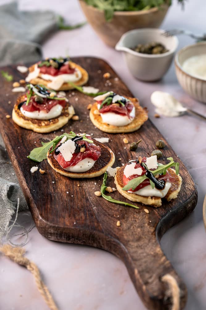 Blini’s met carpaccio, cottage cheese en rucola op kleine pannenkoekjes, geserveerd als feestelijk hapje.