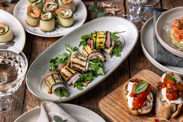 Feestelijk recept voor courgetterolletjes met ricotta, prosciutto, pijnboompitten en rucola; een licht en luxe borrelhapje of voorgerecht dat makkelijk vooraf te maken is.